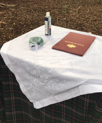 Signing Table with
Scottish National Tartan undercloth and white
on white embroidered linen topper cloth.
Maroon marriage register, green and white
handfasting cords, and a bottle of hand
sanitizer on the table.