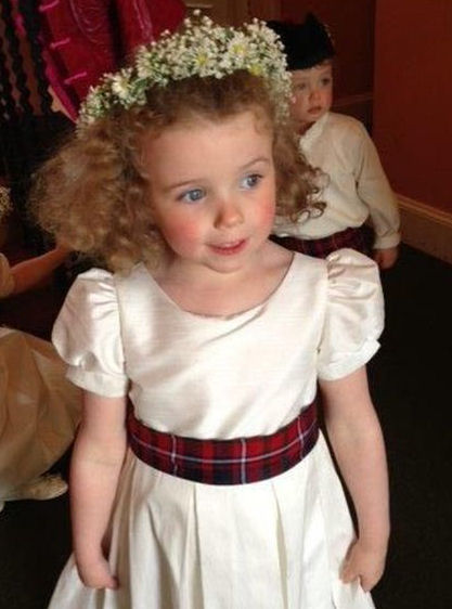 A flower girl wearing a white dress with a
tartan sash and a wreath of baby's breath on her
head