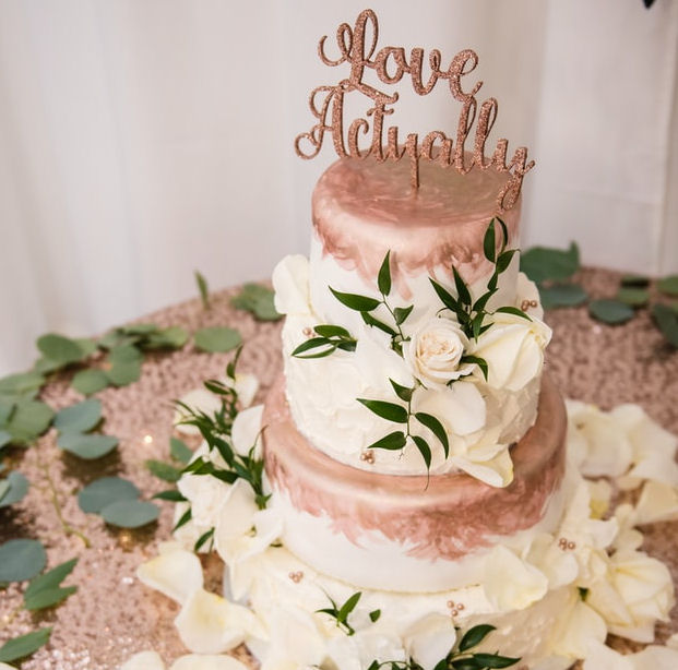 Wedding Cake with Love Actually topper
standing on a table covered in white rose petals
and green leaves