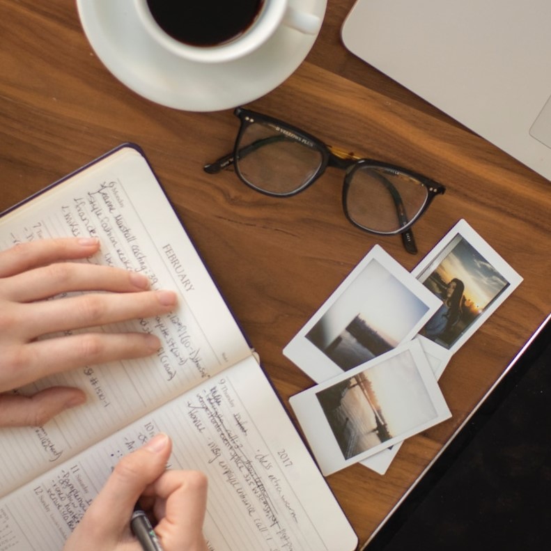 Hands
holding a pen and writing in a notebook. A cup of
coffee, a pair of eyeglasses and some photos are
nearby
